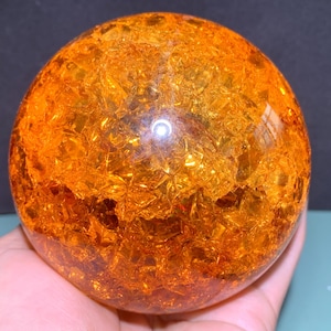 Könnte beinhalten: Eine dekorative orangefarbene Glaskugel mit einer rissigen, strukturierten Oberfläche. Die Kugel hat eine leuchtende Bernsteinfarbe und reflektiert das Licht, wodurch ein funkelnder Effekt entsteht. Die Kugel wird in einer Hand gehalten.