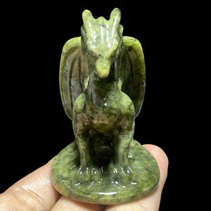Op de afbeelding: Een groene drakenfiguur met gedetailleerde vleugels en een zittende pose. De sculptuur is gemaakt van een gespikkeld groen materiaal en staat op een cirkelvormige basis. De draak heeft een gedetailleerd gezicht en staat tegen een zwarte achtergrond.