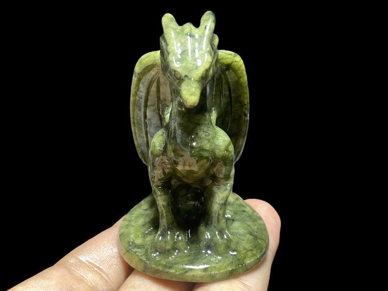 Op de afbeelding: Een groene drakenfiguur met gedetailleerde vleugels en een zittende pose. De sculptuur is gemaakt van een gespikkeld groen materiaal en staat op een cirkelvormige basis. De draak heeft een gedetailleerd gezicht en staat tegen een zwarte achtergrond.