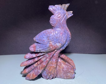 Könnte beinhalten: Eine geschnitzte Steinskulptur eines Vogels mit langem, fließendem Schwanz. Der Vogel ist hauptsächlich lila mit roten und braunen Akzenten. Die Skulptur ist detailliert, mit sichtbaren Federn und einer strukturierten Oberfläche. Der Vogel sitzt auf einem Sockel.