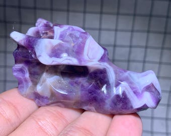 Op de afbeelding: Een gebeeldhouwde drakenhoofdsculptuur van amethist. Het drakenhoofd is een mix van paarse en witte kleuren. De sculptuur wordt in een hand gehouden.