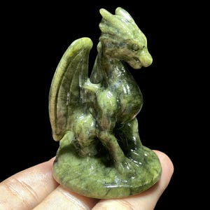 Op de afbeelding: Een gebeeldhouwd groen drakenbeeldje met gedetailleerde vleugels en een getextureerd oppervlak. De draak zit, met zijn hoofd opzij gedraaid. Het beeldje is ongeveer 10 cm hoog.