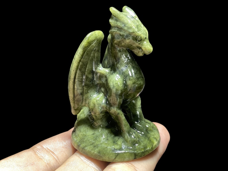 Op de afbeelding: Een gebeeldhouwd groen drakenbeeldje met gedetailleerde vleugels en een getextureerd oppervlak. De draak zit, met zijn hoofd opzij gedraaid. Het beeldje is ongeveer 10 cm hoog.