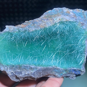 Puede incluir: Una roca verde y gris con una superficie verde grande y lisa. La superficie verde está cubierta de cristales finos, blancos y en forma de aguja.