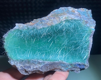 May include: A green and gray rock with a large, smooth, green surface. The green surface is covered in thin, white, needle-like crystals.
