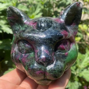 May include: A carved cat head sculpture made of a stone with a mix of colors. The sculpture has a black base color with red and green accents. The cat's features are well-defined, with visible eyes, nose, and ears.