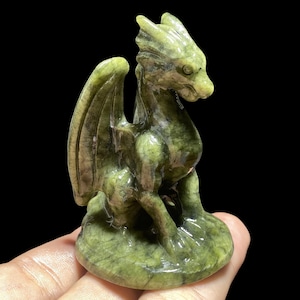 Op de afbeelding: Een groene drakenfiguur met gedetailleerde vleugels en een getextureerd oppervlak. De draak zit op een ronde basis, met zijn hoofd opzij gedraaid. Het beeldje is gemaakt van een steenachtig materiaal.