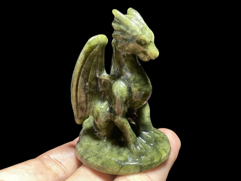 Op de afbeelding: Een groene drakenfiguur met gedetailleerde vleugels en een getextureerd oppervlak. De draak zit op een ronde basis, met zijn hoofd opzij gedraaid. Het beeldje is gemaakt van een steenachtig materiaal.