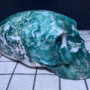May include: A carved skull made of green and white stone. The skull has detailed features, including eye sockets, a nose cavity, and teeth. The stone has a marbled appearance with swirls of green and white. The skull is resting on a checkered surface.