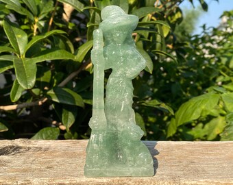 May include: A translucent green crystal figurine of a woman wearing a hat and holding a staff. The sculpture is set against a backdrop of green foliage and rests on a wooden surface. The figurine is approximately 6 inches tall.