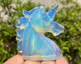 May include: A translucent, opalescent unicorn figurine. The sculpture is carved from a light blue and white material, with a prominent horn and flowing mane. The figurine is held in a hand, with a blurred green background.