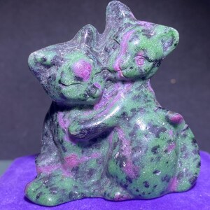 May include: A green and pink carved stone sculpture of two cats embracing. The cats are made of a mottled green stone with pink accents.