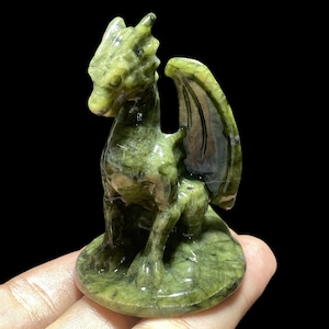 Op de afbeelding: Een gebeeldhouwd groen drakenbeeldje met gedetailleerde vleugels en een zittende pose. De draak is gemaakt van een gespikkelde groene steen en staat op een ronde basis. Het beeldje staat tegen een zwarte achtergrond.