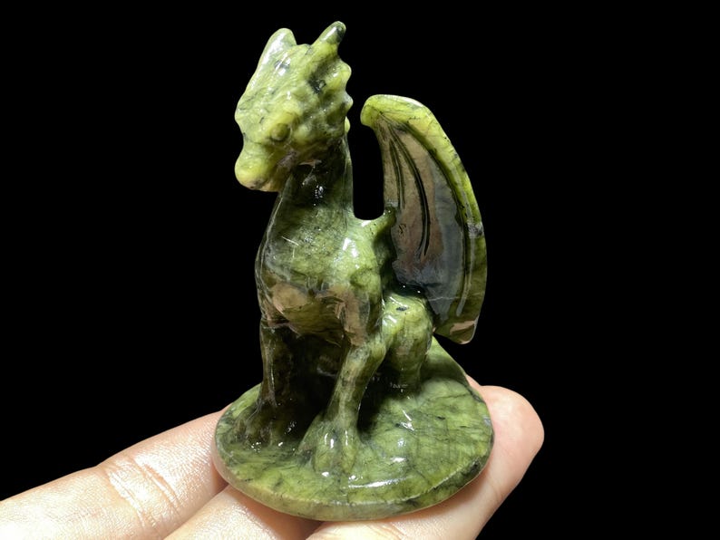 Op de afbeelding: Een gebeeldhouwd groen drakenbeeldje met gedetailleerde vleugels en een zittende pose. De draak is gemaakt van een gespikkelde groene steen en staat op een ronde basis. Het beeldje staat tegen een zwarte achtergrond.