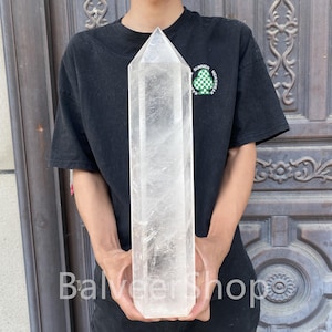May include: A large, clear quartz crystal obelisk. The crystal is held upright, showcasing its pointed top and rectangular shape. The obelisk is transparent with visible internal patterns.