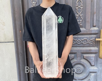 Op de afbeelding: Een grote, heldere kwartskristal obelisk. Het kristal wordt rechtop gehouden, waarbij de puntige top en rechthoekige vorm worden getoond. De obelisk is transparant met zichtbare interne patronen.