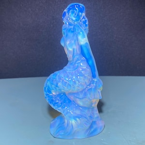 May include: A blue opalescent glass mermaid figurine with a detailed design. The mermaid is sitting on a wave with her tail curled around her.