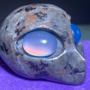 May include: A grey stone sculpture of an alien head with two blue eyes. The eyes are made of glass and have an iridescent finish.