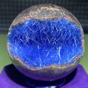Op de afbeelding: Een geode met een donkerbruine buitenkant en een helderblauwe binnenkant gevuld met kristallen. De geode ligt op een paarse ondergrond.
