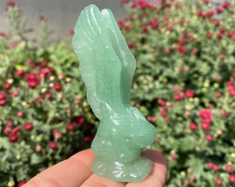 May include: A light green crystal sculpture of a rabbit with detailed carved features. The bunny figurine is standing upright with its ears raised. The sculpture is held in a hand, with a background of red flowers and green foliage.