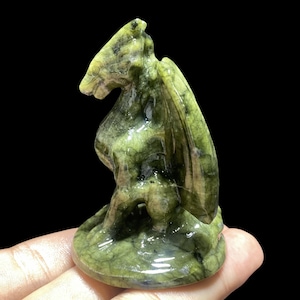 Op de afbeelding: Een groene en bruine gebeeldhouwde stenen drakenfiguur. De draak heeft vleugels en zit op een ronde basis. Het beeldje wordt in een hand gehouden tegen een zwarte achtergrond.