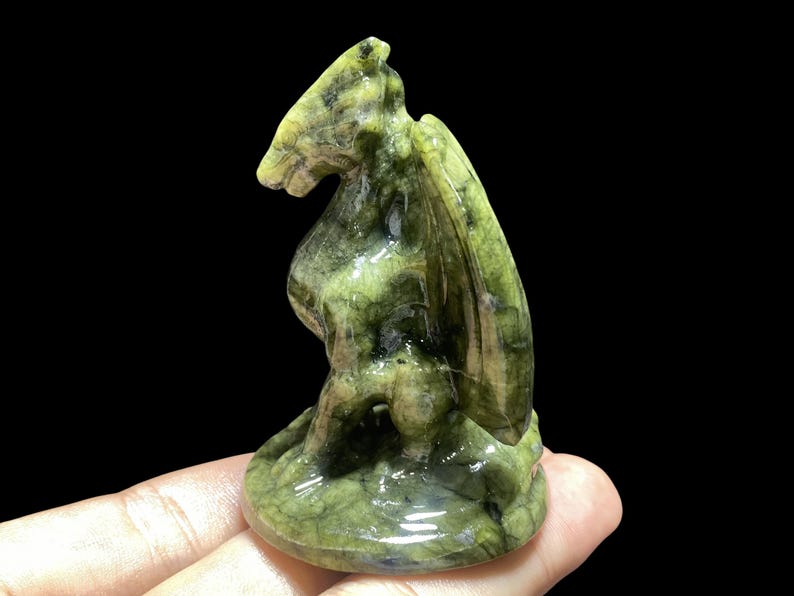 Op de afbeelding: Een groene en bruine gebeeldhouwde stenen drakenfiguur. De draak heeft vleugels en zit op een ronde basis. Het beeldje wordt in een hand gehouden tegen een zwarte achtergrond.