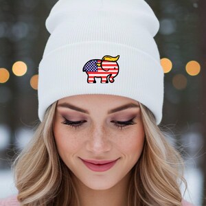 May include: A white beanie hat with a cartoon elephant design incorporating the American flag and yellow hair. The hat is made of a soft, ribbed knit material. The hat is worn by a person with blonde hair.