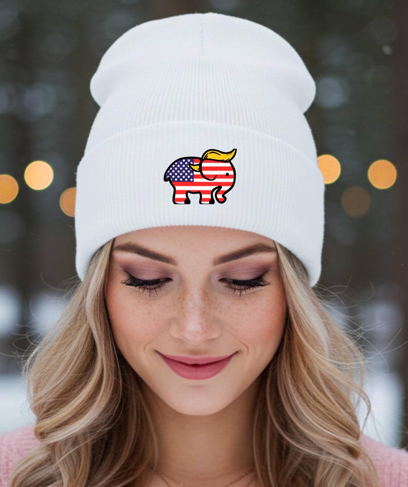 May include: A white beanie hat with a cartoon elephant design incorporating the American flag and yellow hair. The hat is made of a soft, ribbed knit material. The hat is worn by a person with blonde hair.