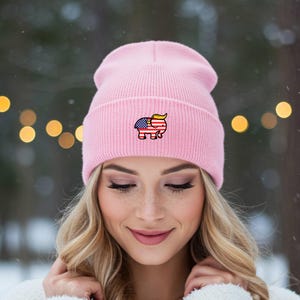May include: A pale pink beanie hat with a ribbed knit design. The hat has a small embroidered elephant motif, incorporating the American flag and a yellow hairstyle. The hat is worn by a person with blonde hair.