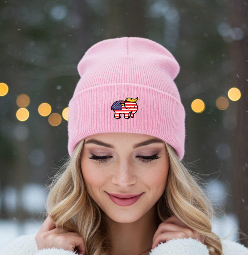 May include: A pale pink beanie hat with a ribbed knit design. The hat has a small embroidered elephant motif, incorporating the American flag and a yellow hairstyle. The hat is worn by a person with blonde hair.