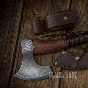 May include: A handcrafted axe with a patterned steel blade and a wooden handle. The handle is wrapped in leather and includes a leather sheath and belt loop. The axe is displayed on a wooden surface. The text "BLADE FLARE" is visible.