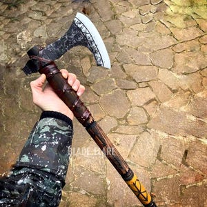 Könnte beinhalten: Eine schwarze und silberne Axt mit einem Holzstiel, der in braunes Leder gewickelt ist. Der Stiel hat eine gelbe Lederumwicklung mit einem Muster. Die Axt wird von einer Hand gehalten, die eine Tarnjacke trägt.