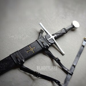 Pode incluir: Uma espada medieval com uma lâmina prateada e um cabo preto, numa bainha preta. A bainha apresenta um emblema de cruz dourada e correias de couro preto. A palavra "BLADEFLARE" é visível.