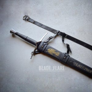 Puede incluir: Una vaina de cuero negro con detalles dorados y la inscripción "Lux Vincit Tenebras" se muestra con una espada plateada unida. La vaina tiene una correa y una hebilla para llevar.