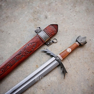 May include: A medieval sword with a wooden handle and a silver blade, next to a red leather scabbard. The scabbard is decorated with carved patterns and metal accents. The sword's hilt has a shield-shaped pommel.