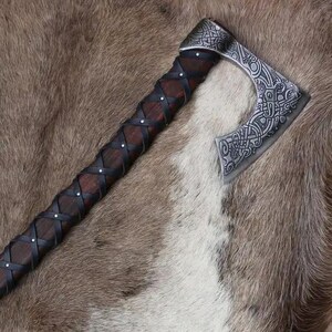 May include: A Viking axe with a dark wooden handle wrapped in black leather and brown leather cross-stitching. The axe head is silver with intricate engraved patterns. The axe is displayed on a brown and white fur-like surface. The text "BLADEFLARE" is visible.
