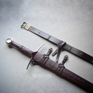 Puede incluir: Una espada de estilo medieval con una vaina y un cinturón de cuero marrón. La espada presenta una hoja plateada y una empuñadura decorativa. La vaina está adornada con patrones en relieve y detalles metálicos. El cinturón tiene cierres metálicos decorativos.