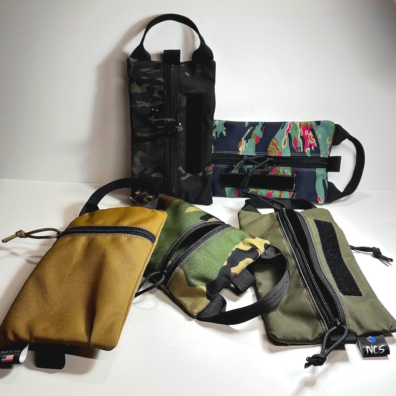 May include: An assortment of zippered pouches in various camouflage and solid colors, including black, olive green, tan, and a patterned blue and pink design. Each pouch features a black zipper, a handle, and a small Velcro patch. Some have a drawstring.