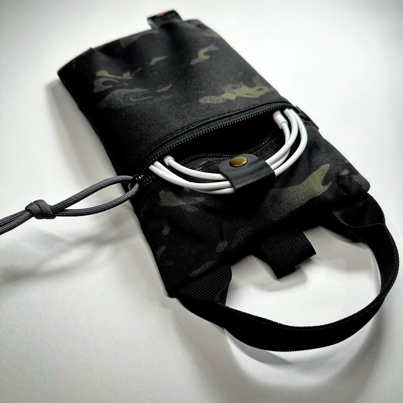 May include: Black camouflage pouch with a zipper closure, a black strap, and a gray cord. The pouch contains white charging cables. The fabric has a pattern of black, green, and brown. The pouch is designed to hold small electronics.