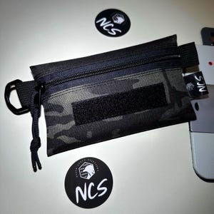 May include: Black and gray camouflage pouch with a black zipper and a Velcro patch. The pouch has a black carabiner clip and a small tag with the letters "NCS". Two black and white circular stickers with the text "NCS" are also visible.