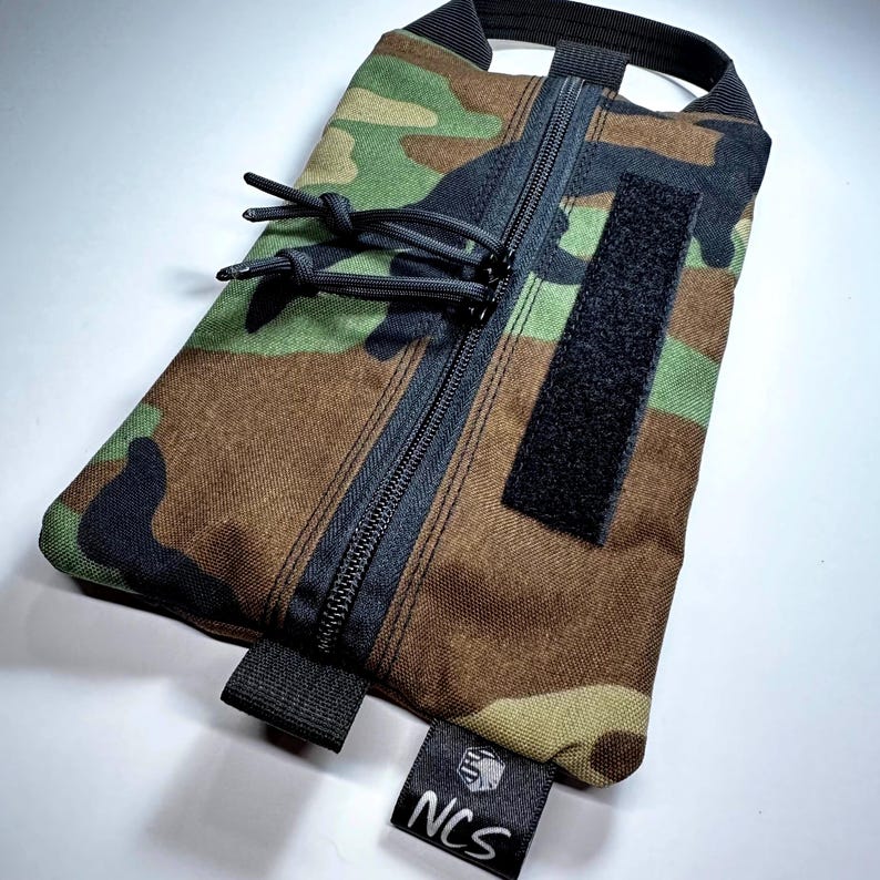 May include: Camouflage pattern pouch with a black zipper, pull cords, and a Velcro patch. The pouch has a black handle and a small tag with the letters "NCS". The color scheme includes shades of green, brown, and black.