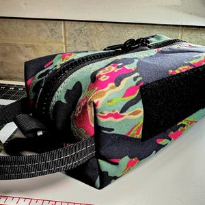 May include: A rectangular camouflage-patterned pouch with a black zipper, handle, and Velcro patch. The fabric features a mix of dark blue, teal, pink, and brown colors. The pouch is designed with a sturdy construction and a practical design.