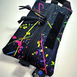 May include: Black rectangular pouch with a vibrant splatter paint design in neon pink, yellow, green, and purple. Features a black zipper, a Velcro patch, and a small handle. The pouch has a tag that reads "NCS".