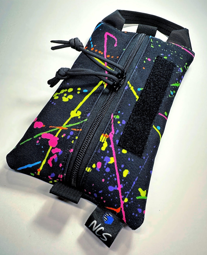 May include: Black rectangular pouch with a vibrant splatter paint design in neon pink, yellow, green, and purple. Features a black zipper, a Velcro patch, and a small handle. The pouch has a tag that reads "NCS".