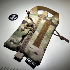May include: A camouflage pattern pouch with a brown zipper and a loop handle. The pouch has a brown velcro patch and a black tag with the letters "NCS" in white. The pouch is designed for carrying small items.