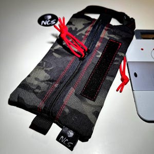May include: A camouflage pattern pouch with red stitching and accents. The pouch has a black zipper, a black Velcro patch, and a black strap. The text "NCS" is visible on a small black tag and a circular sticker.