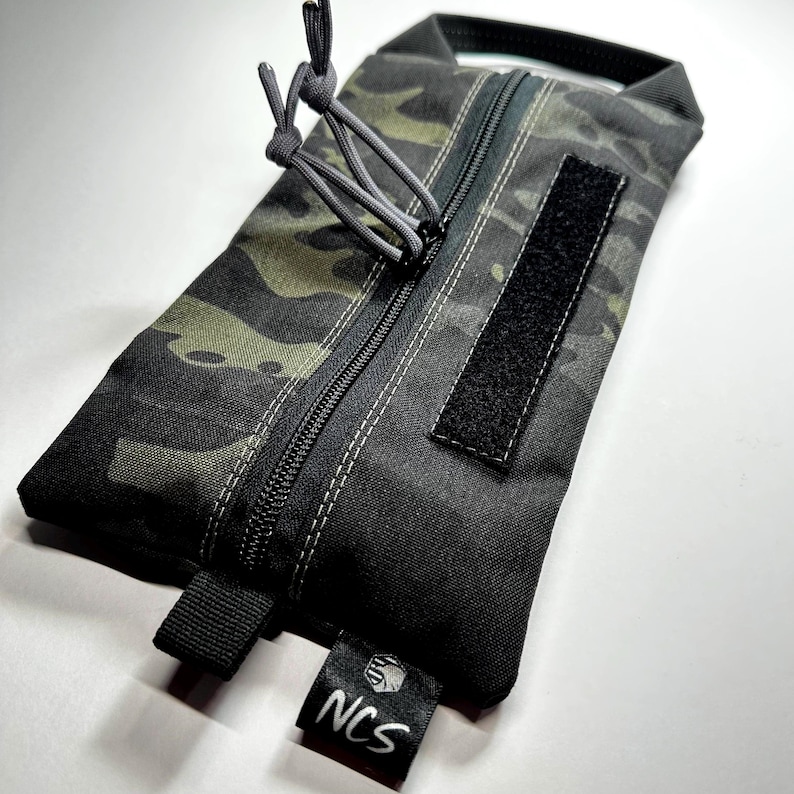 May include: A rectangular camouflage-patterned pouch with a black zipper and a Velcro patch. The pouch has a small black tag with the letters "NCS" printed on it. The pouch is made of durable material and has a gray drawstring.