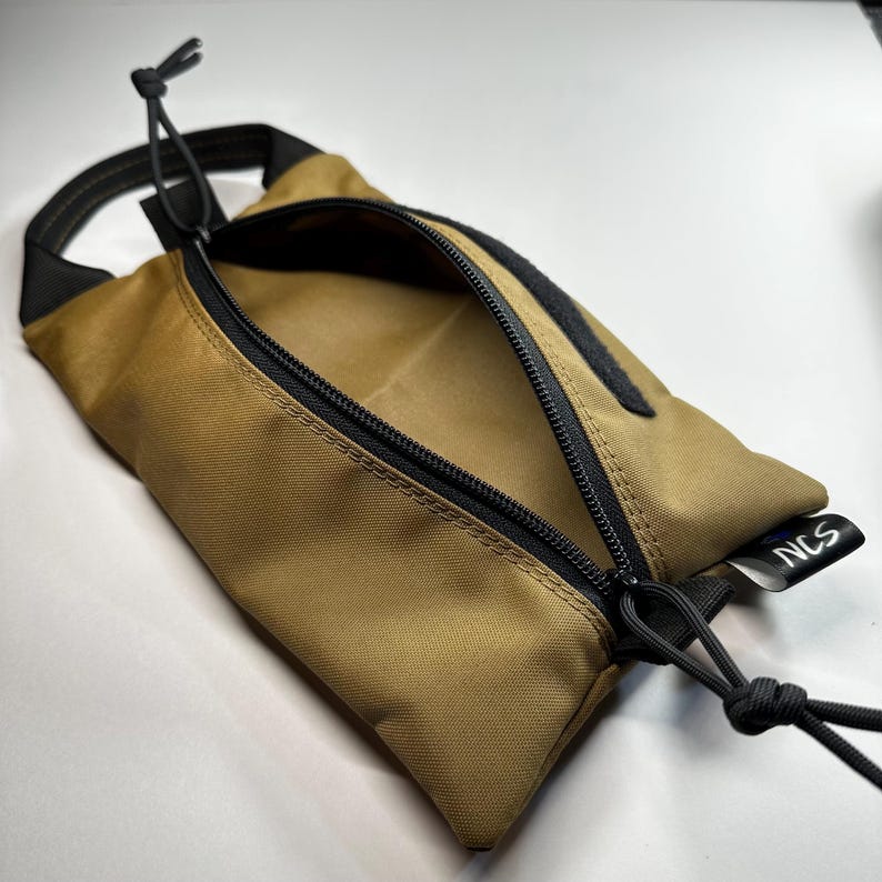 May include: Tan rectangular pouch with a black zipper and black accents. The pouch has a loop handle and a black cord. The text "NCS" is visible on a small tag. The pouch is open, revealing the interior.