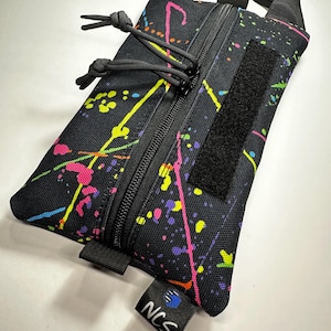 May include: Black rectangular pouch with a colorful splatter paint design in pink, yellow, and blue. The pouch has a black zipper, a Velcro patch, and a small black tag with the letters "NCS". It also has a black handle.