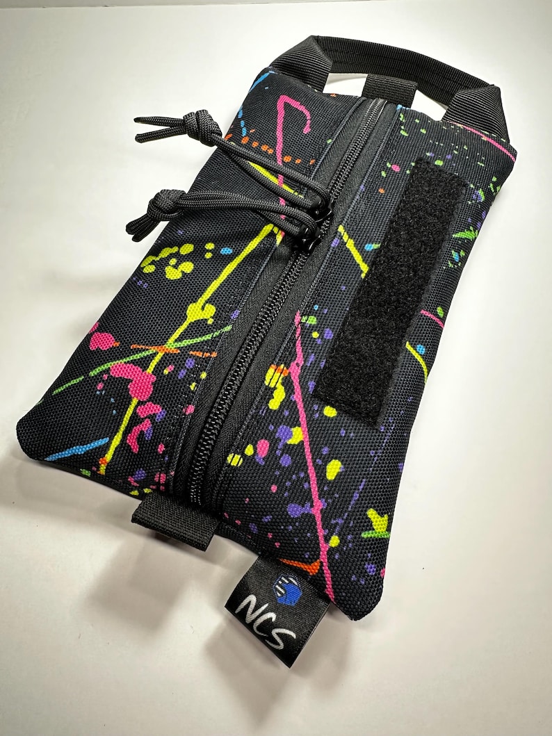 May include: Black rectangular pouch with a colorful splatter paint design in pink, yellow, and blue. The pouch has a black zipper, a Velcro patch, and a small black tag with the letters "NCS". It also has a black handle.